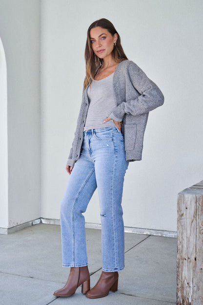 A woman wearing the Cozy Grey Boyfriend Cardigan stands indoors by a wooden bench, looking at the camera.