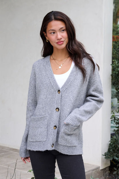 A woman in a Cozy Grey Boyfriend Cardigan, white top, and black pants stands outside, looking to the side with a hand in her pocket.
