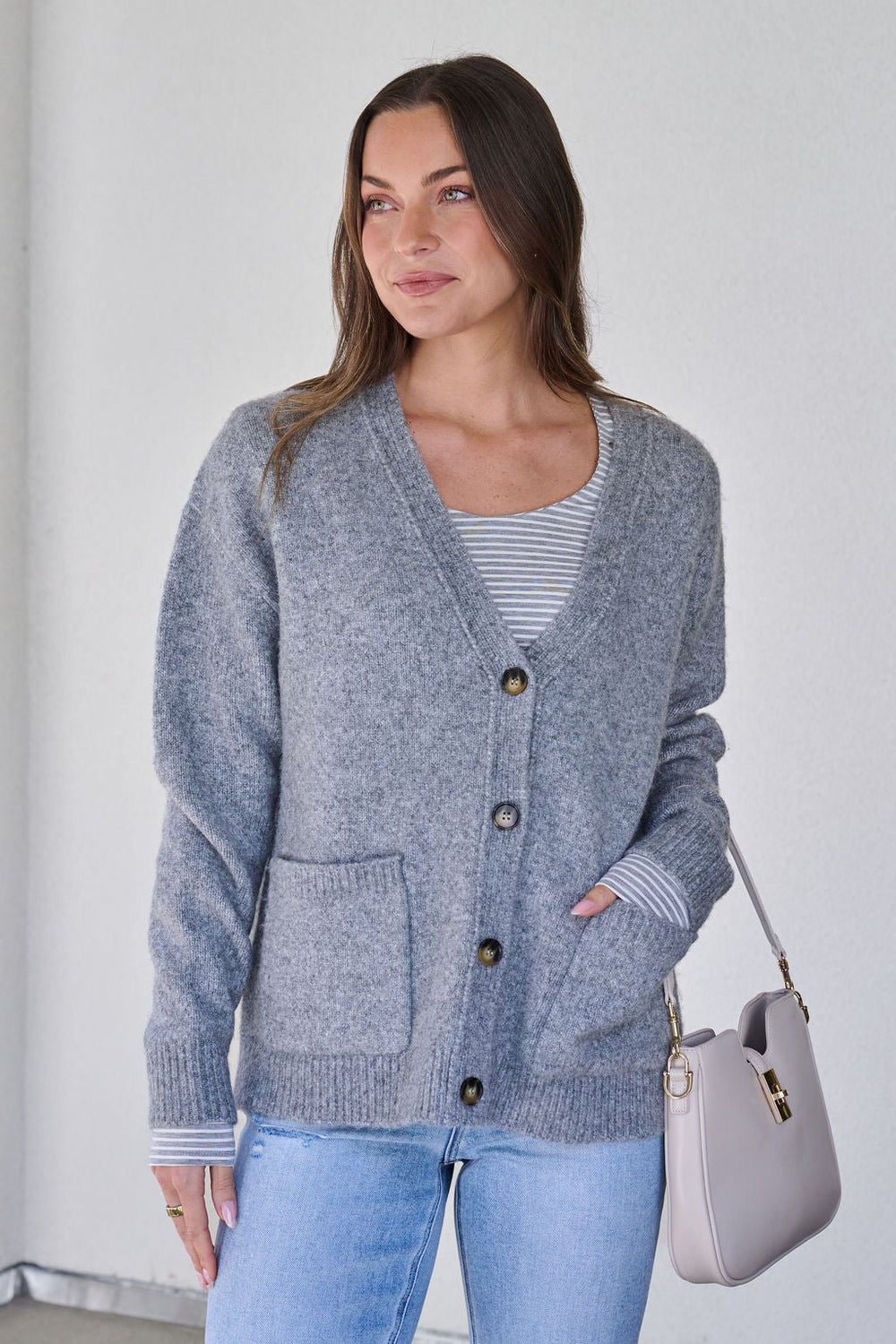 A woman wears the Cozy Grey Boyfriend Cardigan - FINAL SALE over a striped shirt, jeans, and holds a beige handbag against a light background.