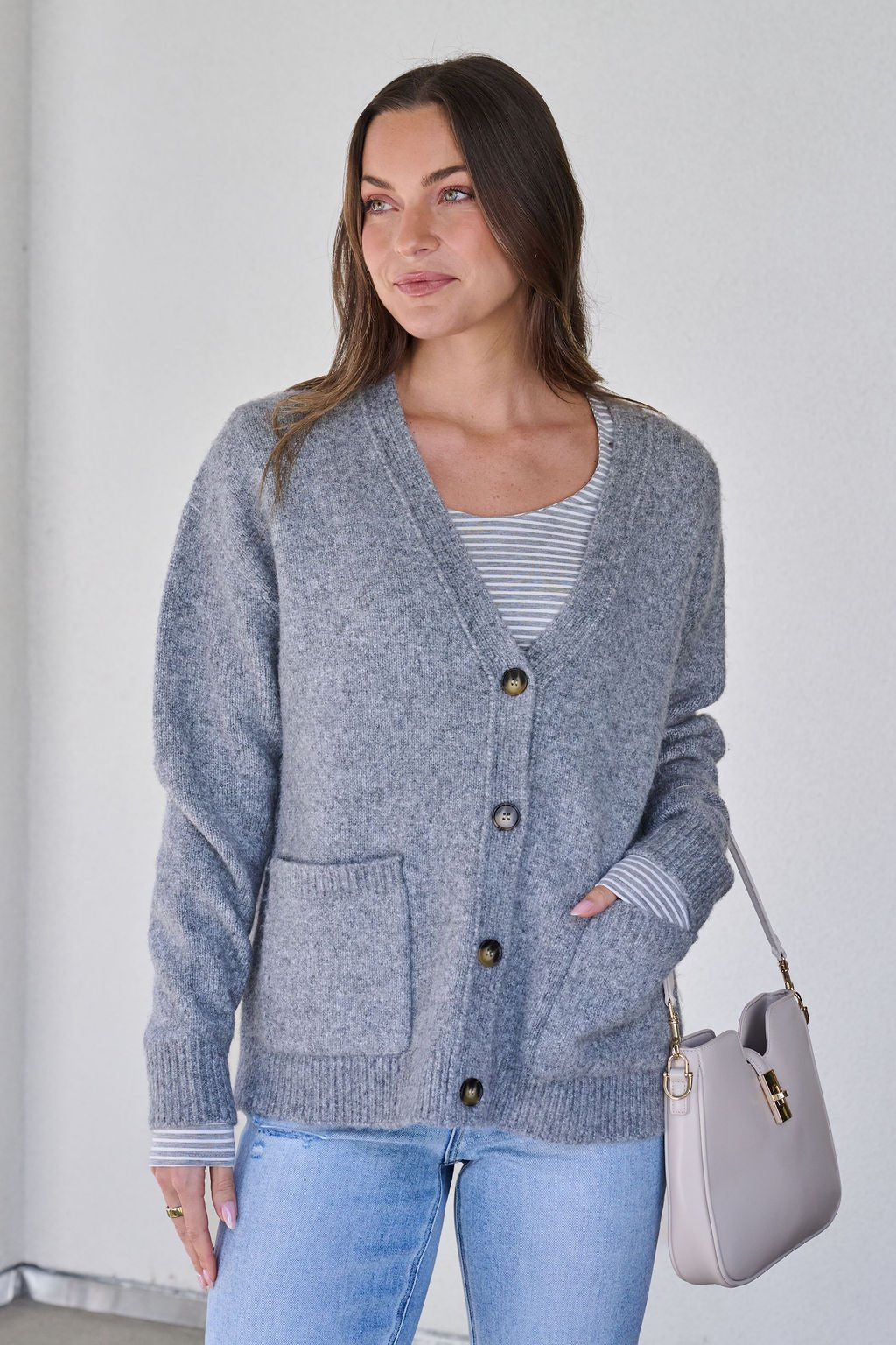 Woman in a Cozy Grey Boyfriend Cardigan over a striped shirt, light blue jeans, holding a beige handbag by a plain wall.