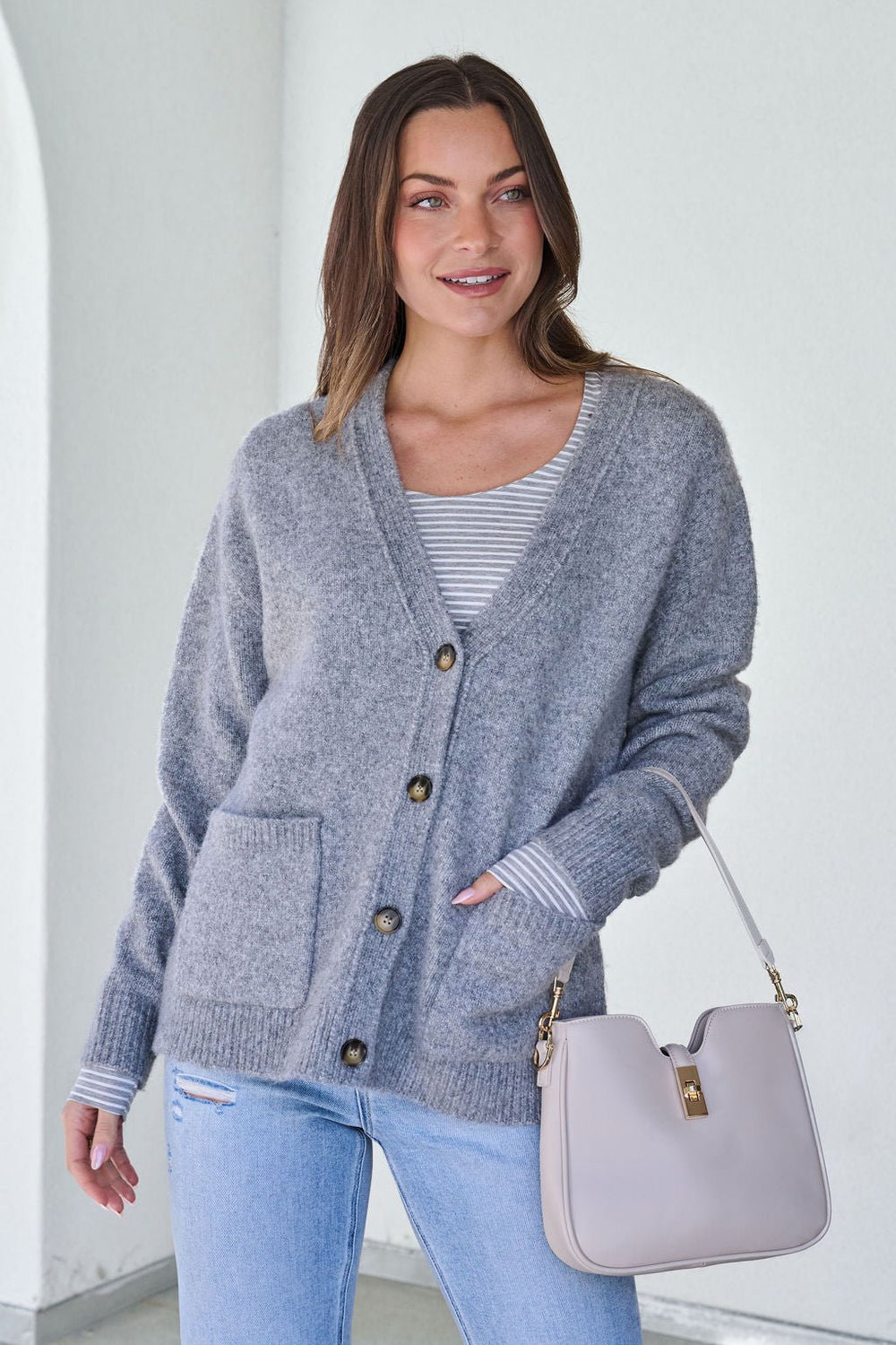 Woman wears the Cozy Grey Boyfriend Cardigan - FINAL SALE over a striped shirt, holding a beige handbag against a white background.