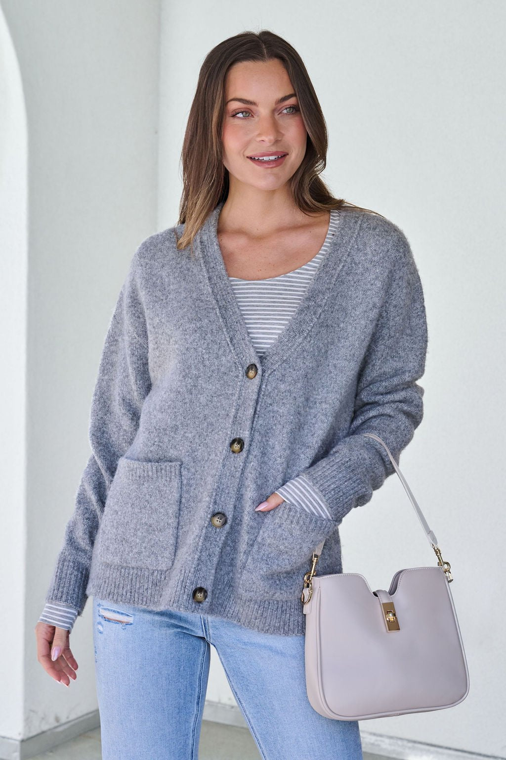 A woman smiles indoors wearing a Cozy Grey Boyfriend Cardigan, striped shirt, light jeans, and holding a light handbag.