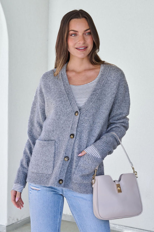 A woman smiles indoors wearing a Cozy Grey Boyfriend Cardigan, striped shirt, light jeans, and holding a light handbag.