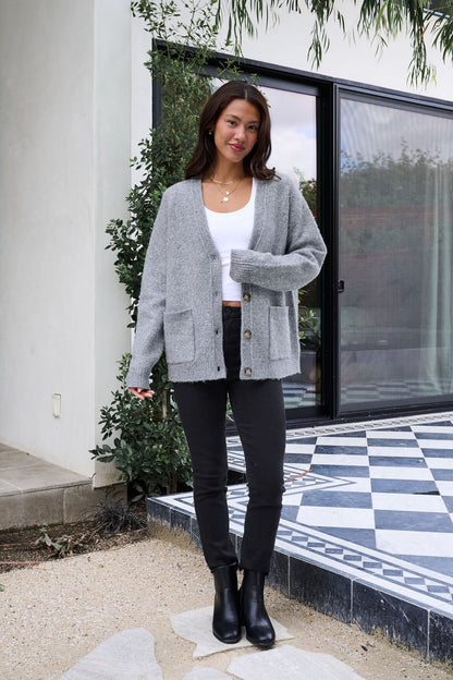 A woman stands on a tiled patio, smiling in the Cozy Grey Boyfriend Cardigan, white top, black pants, and black boots.