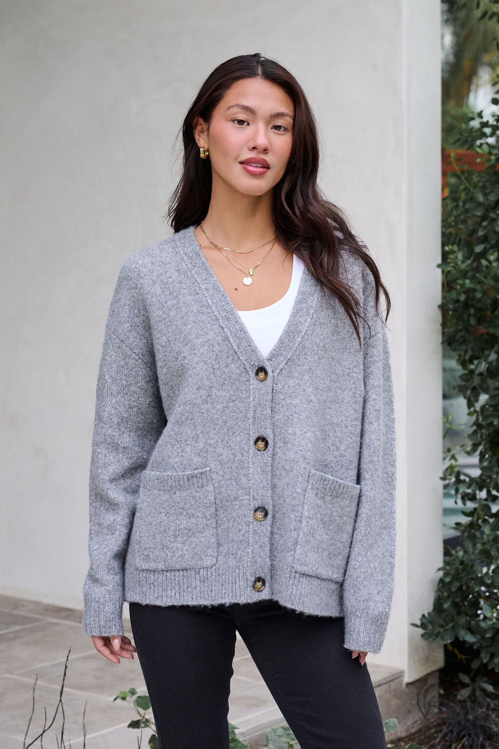 A woman smiles gently at the camera wearing the Cozy Grey Boyfriend Cardigan, a white top, and black pants near greenery.