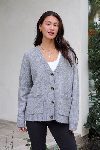 A woman smiles gently at the camera wearing the Cozy Grey Boyfriend Cardigan, a white top, and black pants near greenery.