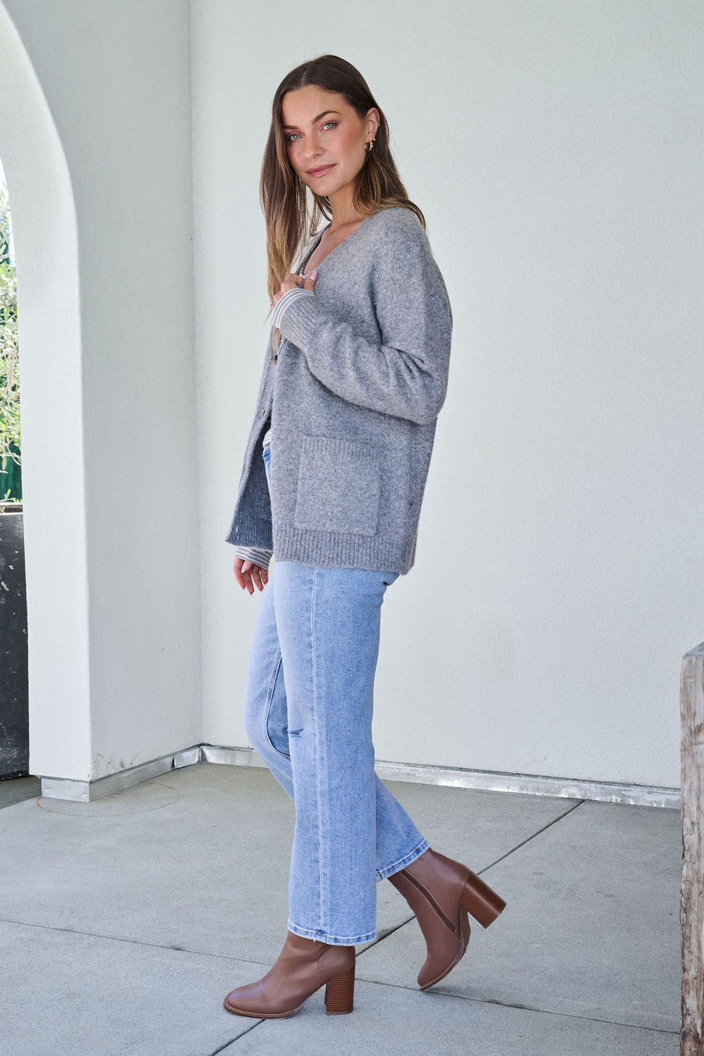 Woman in a Cozy Grey Boyfriend Cardigan - FINAL SALE, light blue jeans, and brown boots stands sideways on a patio.
