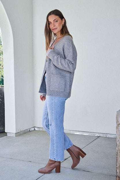 Woman in a Cozy Grey Boyfriend Cardigan, light blue jeans, and brown heeled boots stands sideways on a patio, facing the camera.