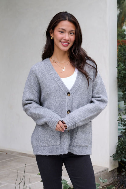 A woman with long dark hair smiles outdoors wearing the Cozy Grey Boyfriend Cardigan over a white top and black pants.