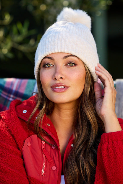 Woman in a red jacket and Cozy Ivory Ribbed Pom Beanie sits outdoors in style, looking at the camera.