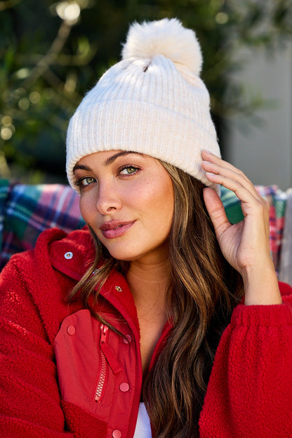 A woman smiles outdoors in a Cozy Ivory Ribbed Pom Beanie, sitting before a blurred green background.