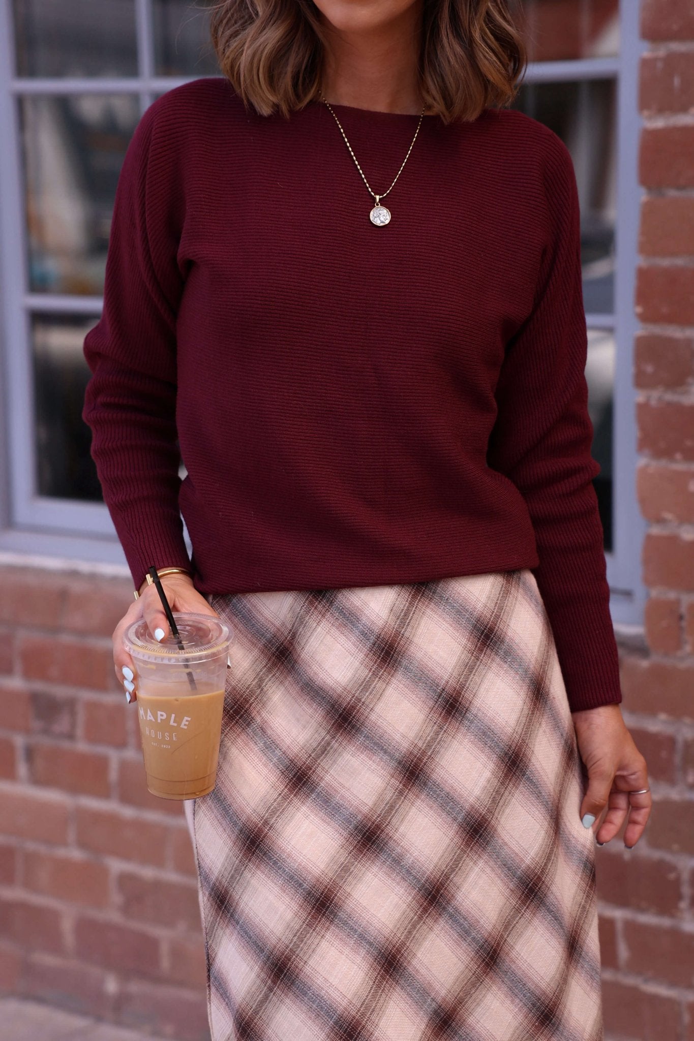Woman in a Cozy Ribbed Boat Neck Sweater - Burgundy, plaid skirt, holding iced coffee in front of a brick wall and window.