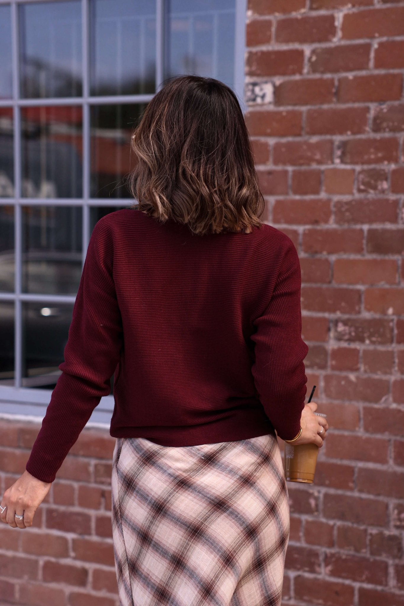 A woman in the Cozy Ribbed Boat Neck Sweater - Burgundy holds an iced drink while facing a brick wall.