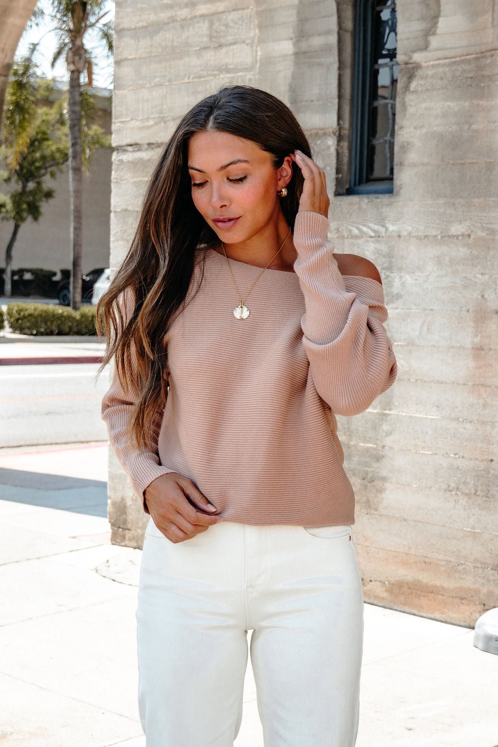 Woman with long brown hair in a Cozy Ribbed Boat Neck Sweater - Camel and white pants stands by a stone building, touching her hair.