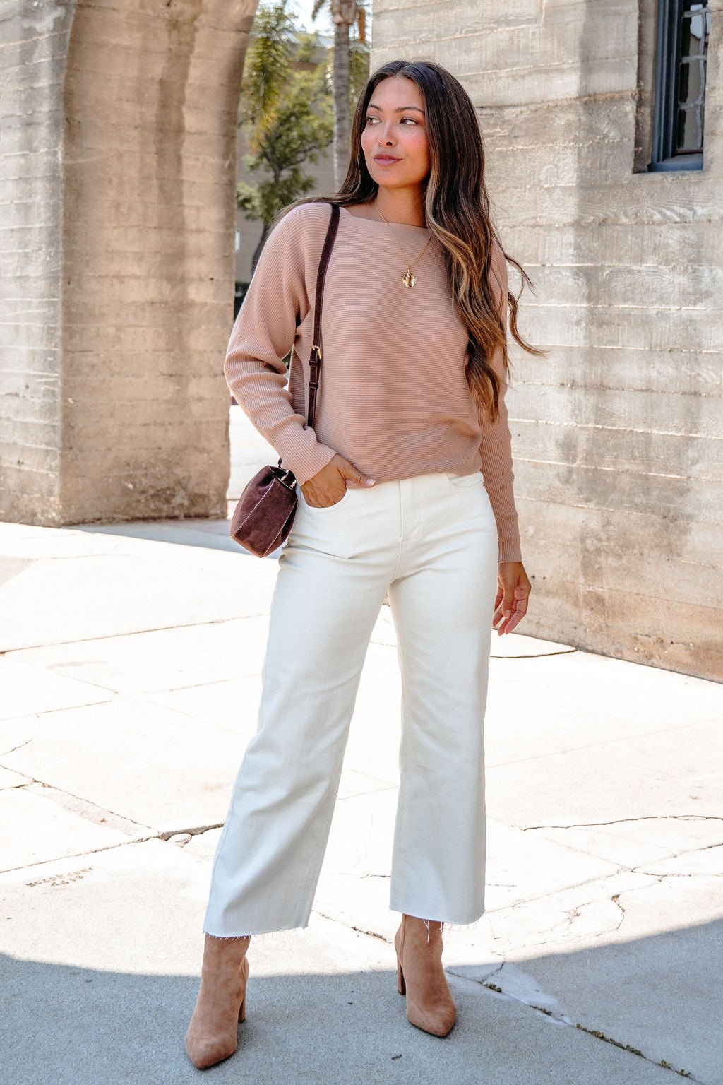 Woman in a Cozy Ribbed Boat Neck Sweater - Camel, white wide-leg pants, brown heels, and a shoulder bag stands outdoors.