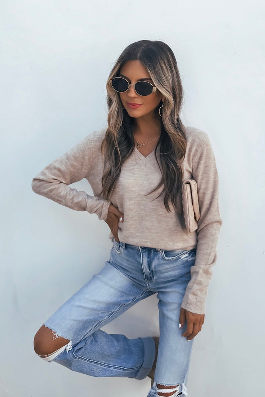 Woman in sunglasses and the Cozy V Neck Sweater - Oatmeal poses against a white wall, wearing ripped jeans and holding a matching clutch.
