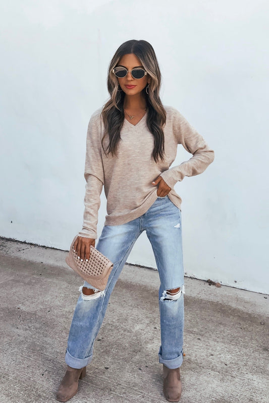 Woman in sunglasses wears the Cozy V Neck Sweater - Oatmeal with ripped jeans, taupe boots, and a matching clutch against a white wall.