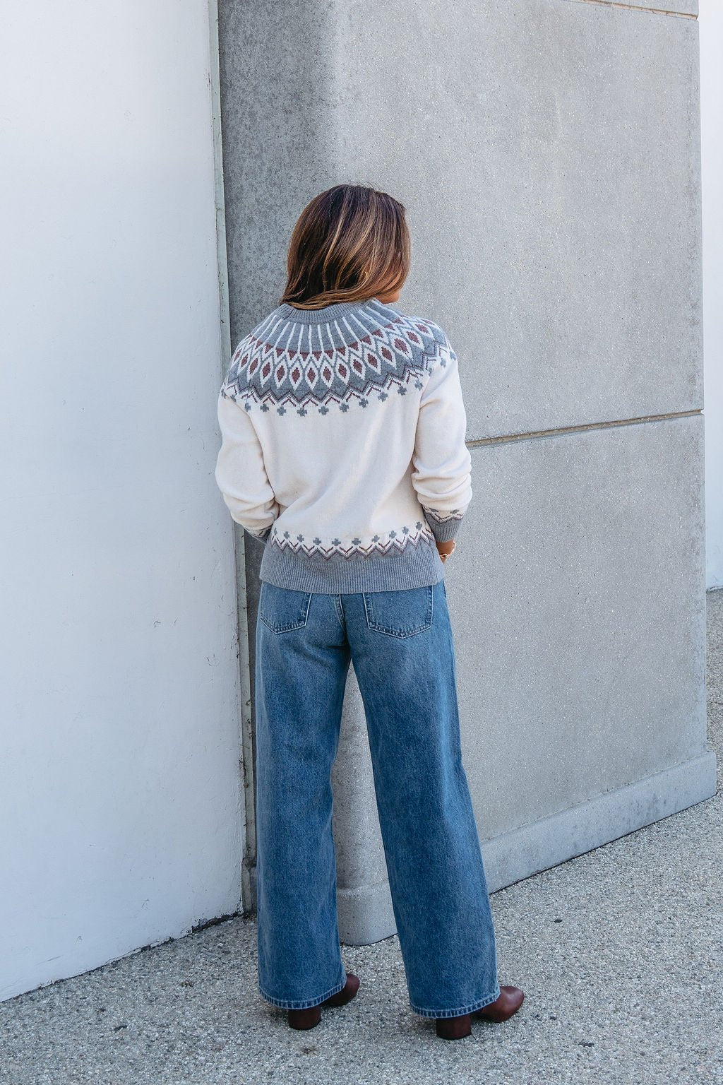 A person in a Cream and Grey Aztec Print Sweater faces a concrete wall—cozy comfort from the autumn collection.