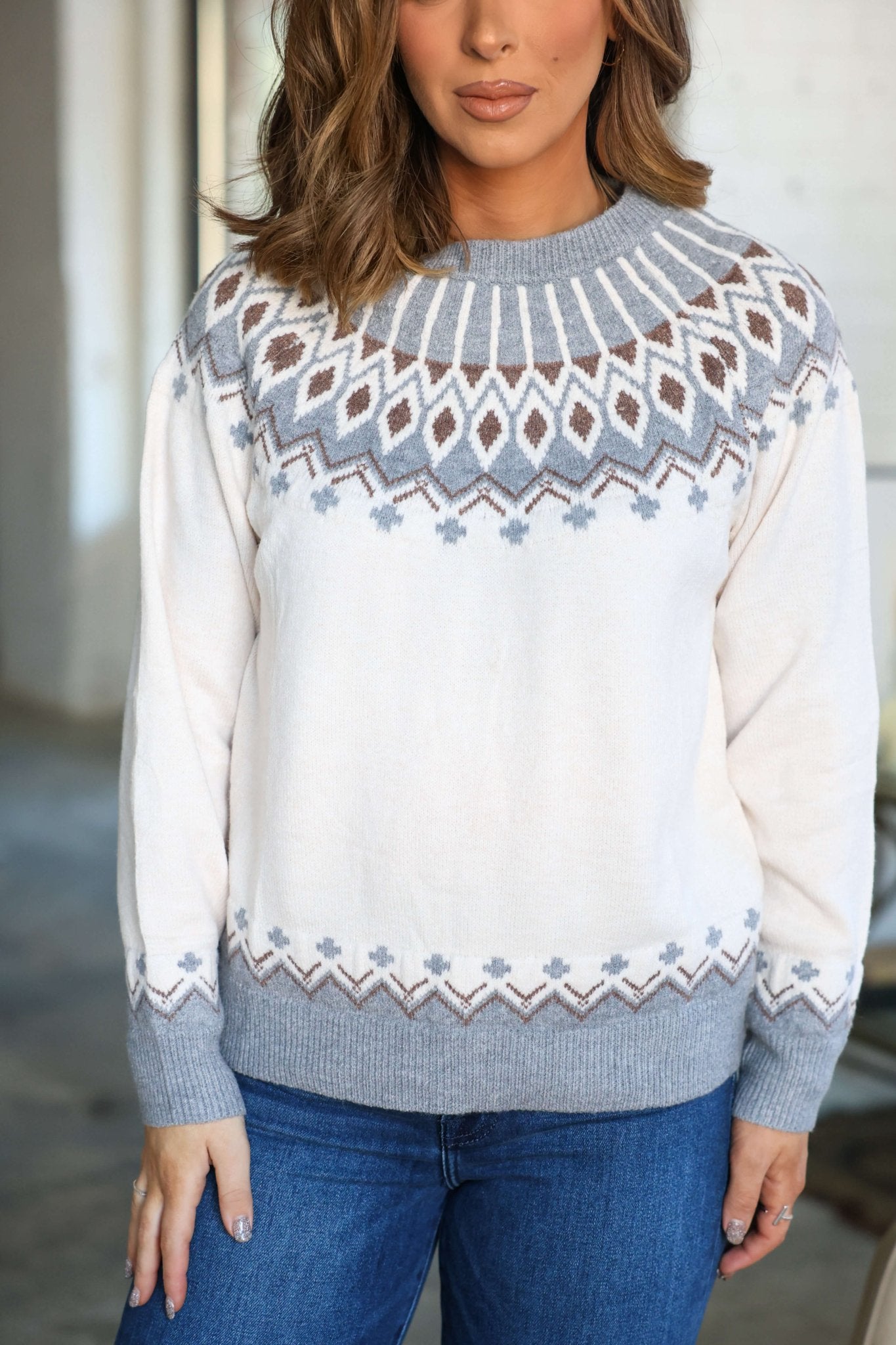 Woman wearing a Cream and Grey Aztec Print Sweater with jeans indoors, her hair styled in loose waves.