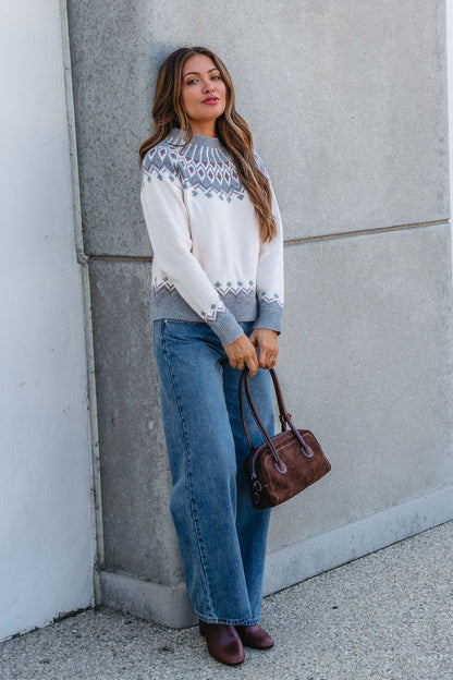 Woman in a Cream and Grey Aztec Print Sweater, wide-leg jeans, and burgundy shoes holds a brown bag by the wall.