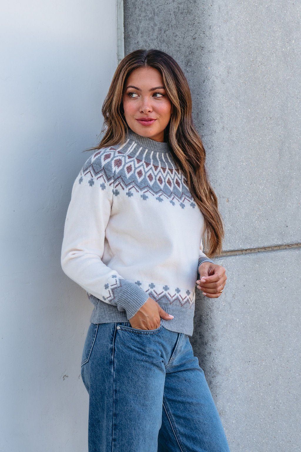 Woman with long brown hair in a Cream and Grey Aztec Print Sweater and blue jeans, standing by a gray concrete wall, radiating comfort.