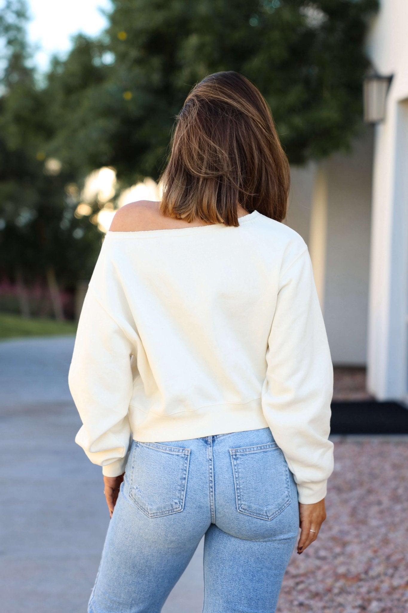 A woman in a Cream Boatneck Lounge Sweatshirt and light blue jeans stands outdoors, back to the camera.