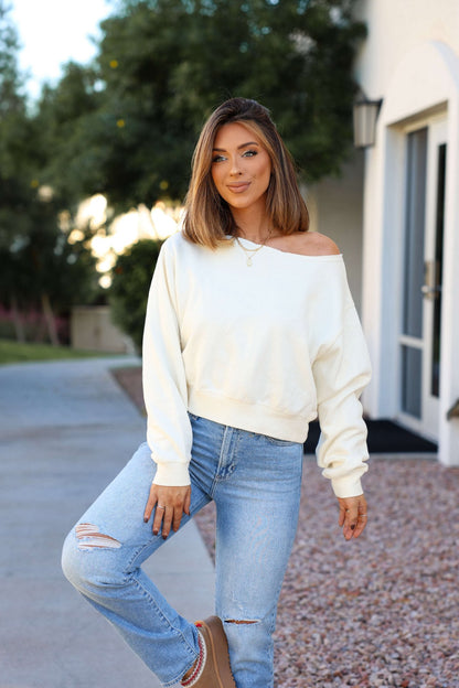 Wearing a Cream Boatneck Lounge Sweatshirt and ripped jeans, a woman smiles outdoors near a building with trees behind her.