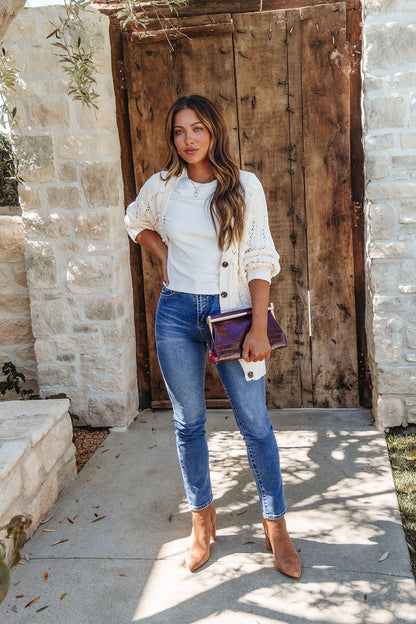 Woman stands by a rustic door in jeans, white top, and our Cream Cable Knit Button Down Cardigan - DOORBUSTER, holding a clutch.