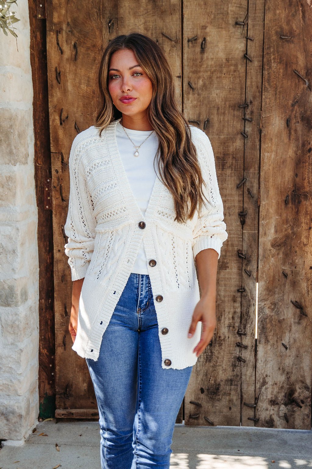 Woman in blue jeans, white top, and Cream Cable Knit Button Down Cardigan - DOORBUSTER stands by a rustic wooden door and stone wall.
