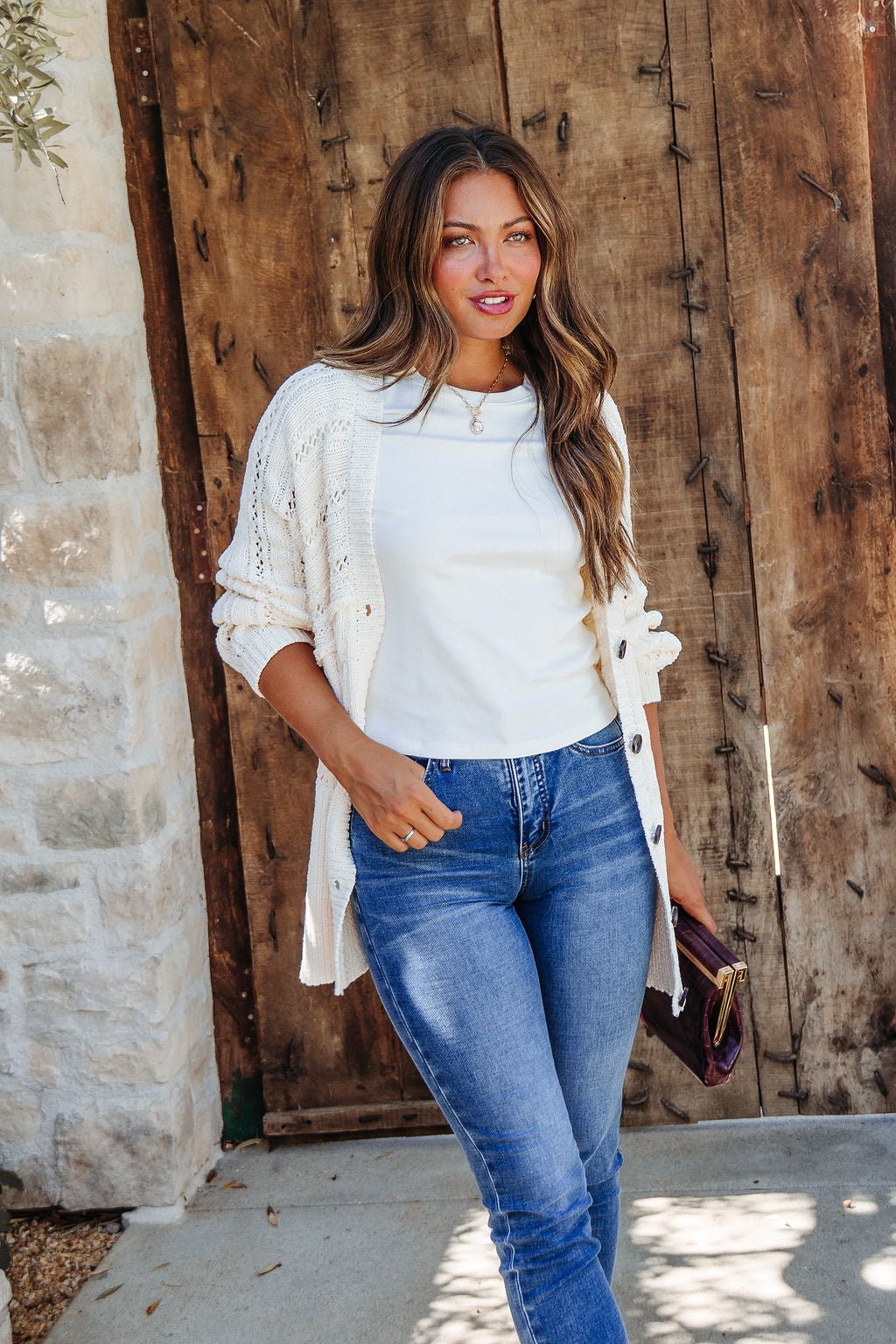 Woman with long hair in the Cream Cable Knit Button Down Cardigan - DOORBUSTER stands by a rustic wooden door, holding a purse.