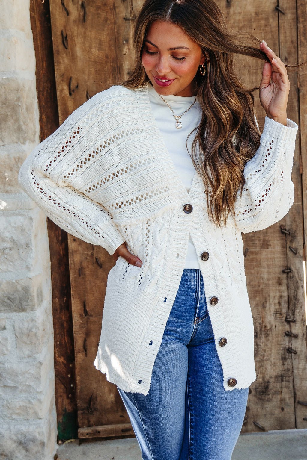 Woman wearing the Cream Cable Knit Button Down Cardigan - DOORBUSTER over a white top and blue jeans, smiling by a rustic door.