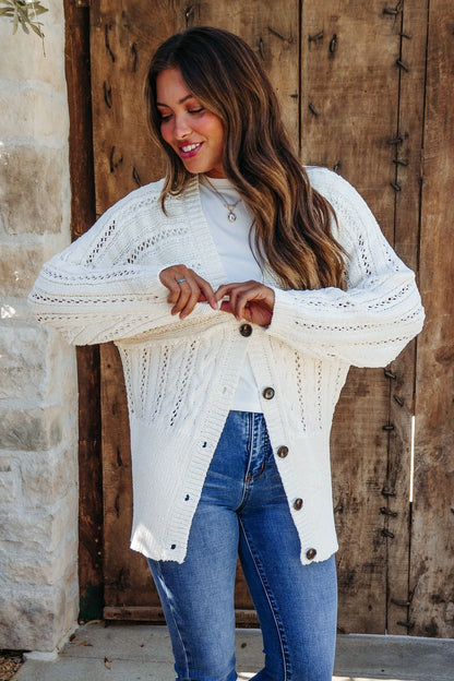Woman with long hair smiles while buttoning the Cream Cable Knit Button Down Cardigan - DOORBUSTER over a white top and jeans.