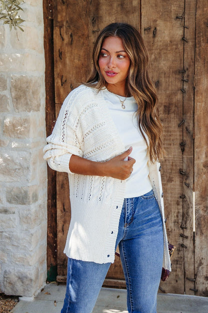 A woman wears the Cream Cable Knit Button Down Cardigan - DOORBUSTER with blue jeans, standing by a rustic wooden door.