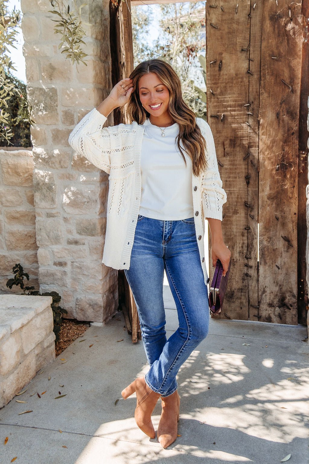Smiling woman wears Cream Cable Knit Button Down Cardigan - DOORBUSTER and blue jeans outside by a wooden gate, holding a clutch. Magnolia exclusive.