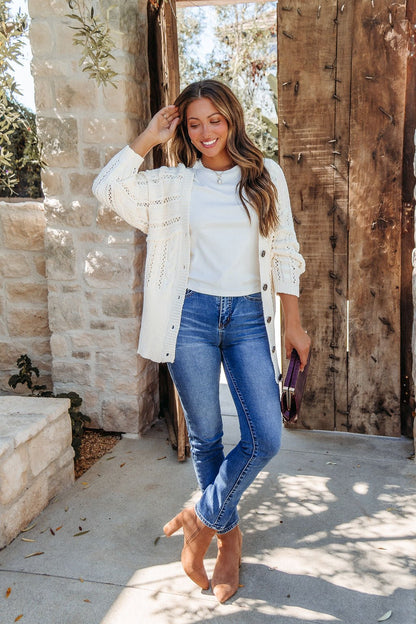 Smiling woman wears Cream Cable Knit Button Down Cardigan - DOORBUSTER and blue jeans outside by a wooden gate, holding a clutch. Magnolia exclusive.