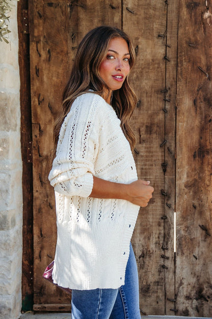 Woman with long wavy hair wears the Cream Cable Knit Button Down Cardigan - DOORBUSTER, posing by a rustic wooden door.