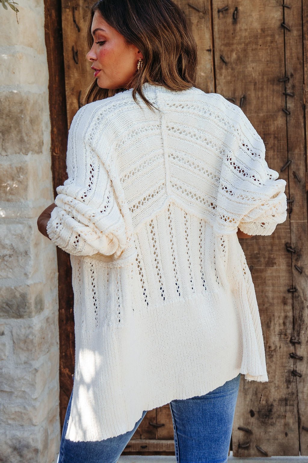 Woman with wavy hair in a Cream Cable Knit Button Down Cardigan - DOORBUSTER and blue jeans, by a rustic wooden door.