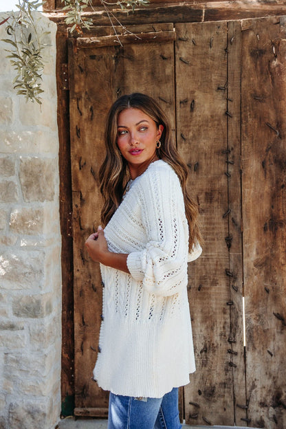 Woman with long brown hair wears the Cream Cable Knit Button Down Cardigan - DOORBUSTER and blue jeans by a rustic wooden door.