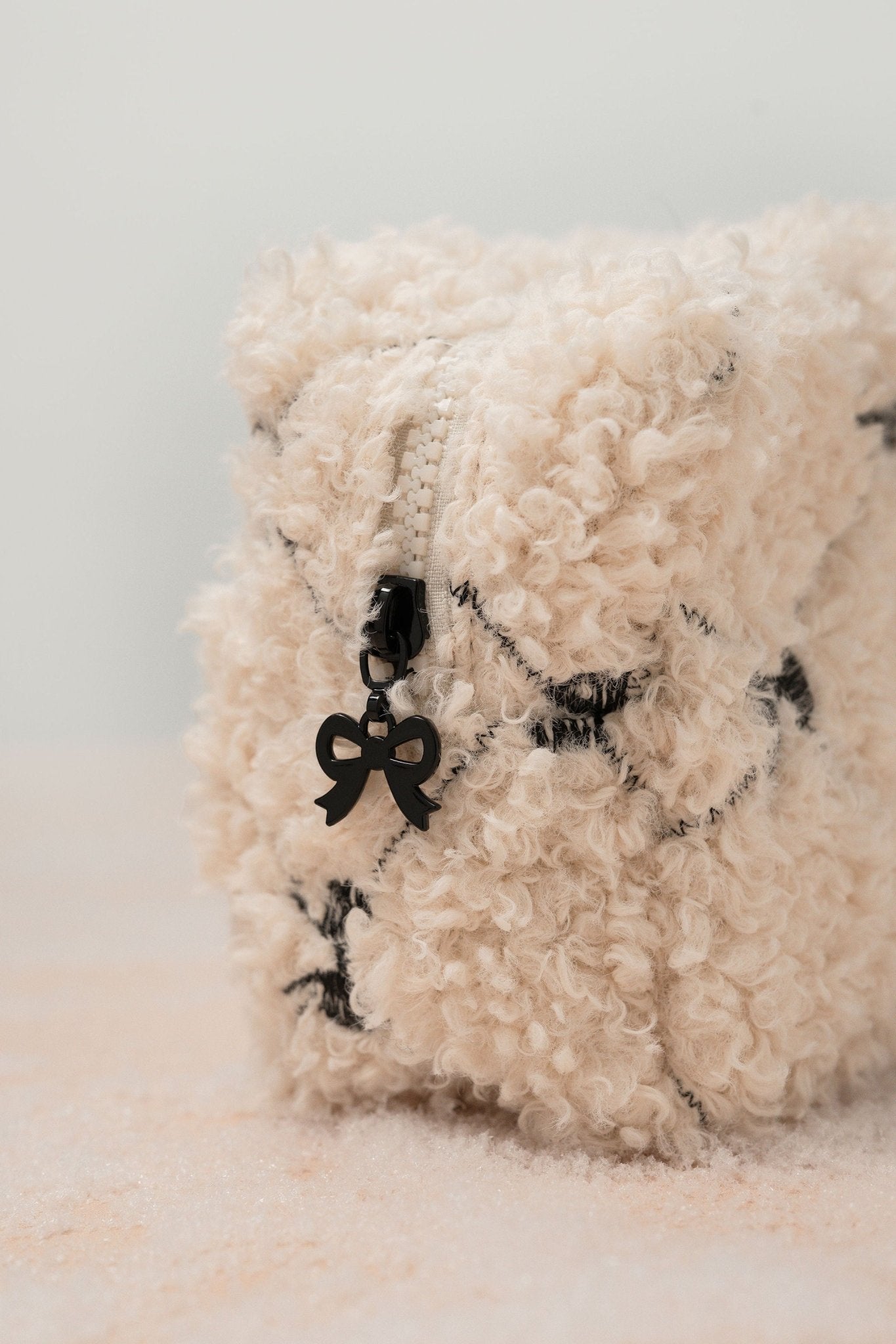 A close-up of the Cream Cherie Sherpa Cosmetic Pouch Bag featuring a zipper and a black bow-shaped pull.