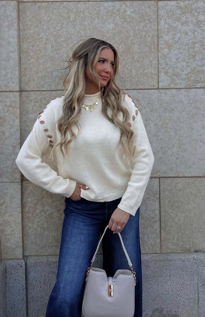 Woman in a Cream Contrast Whipstitch Sweater and blue jeans, holding a beige handbag against a beige tiled wall.