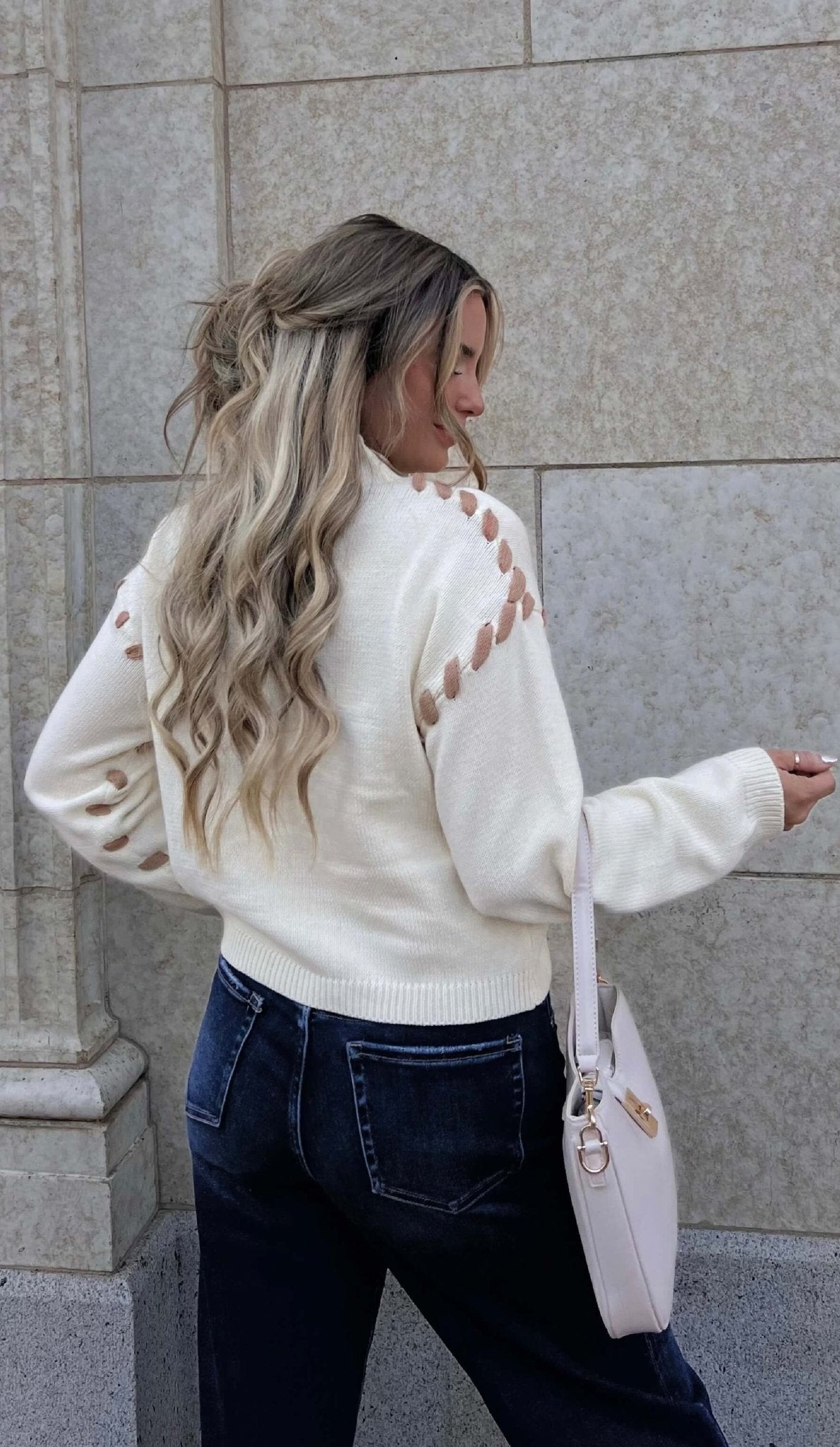 A woman with wavy blonde hair wears the Cream Contrast Whipstitch Sweater and dark jeans, standing sideways by a stone wall.
