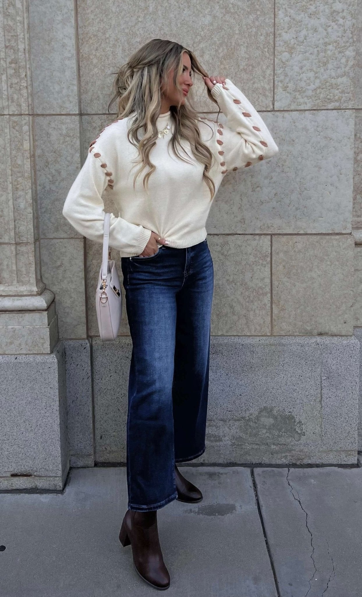 Woman with long wavy hair wears a Cream Contrast Whipstitch Sweater, dark jeans, brown boots, and a pink bag by a stone wall.