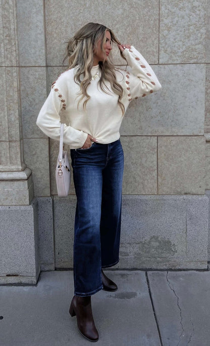 Woman with long wavy hair wears a Cream Contrast Whipstitch Sweater, dark jeans, brown boots, and a pink bag by a stone wall.
