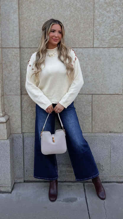 Woman with long blonde hair wears a Cream Contrast Whipstitch Sweater, wide-leg jeans, and brown boots against a stone wall.