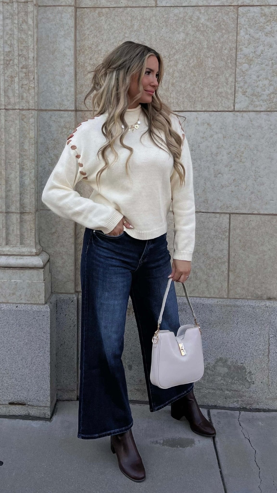 Woman with long wavy hair wears the Cream Contrast Whipstitch Sweater and dark jeans, holding a pale handbag by a stone wall.