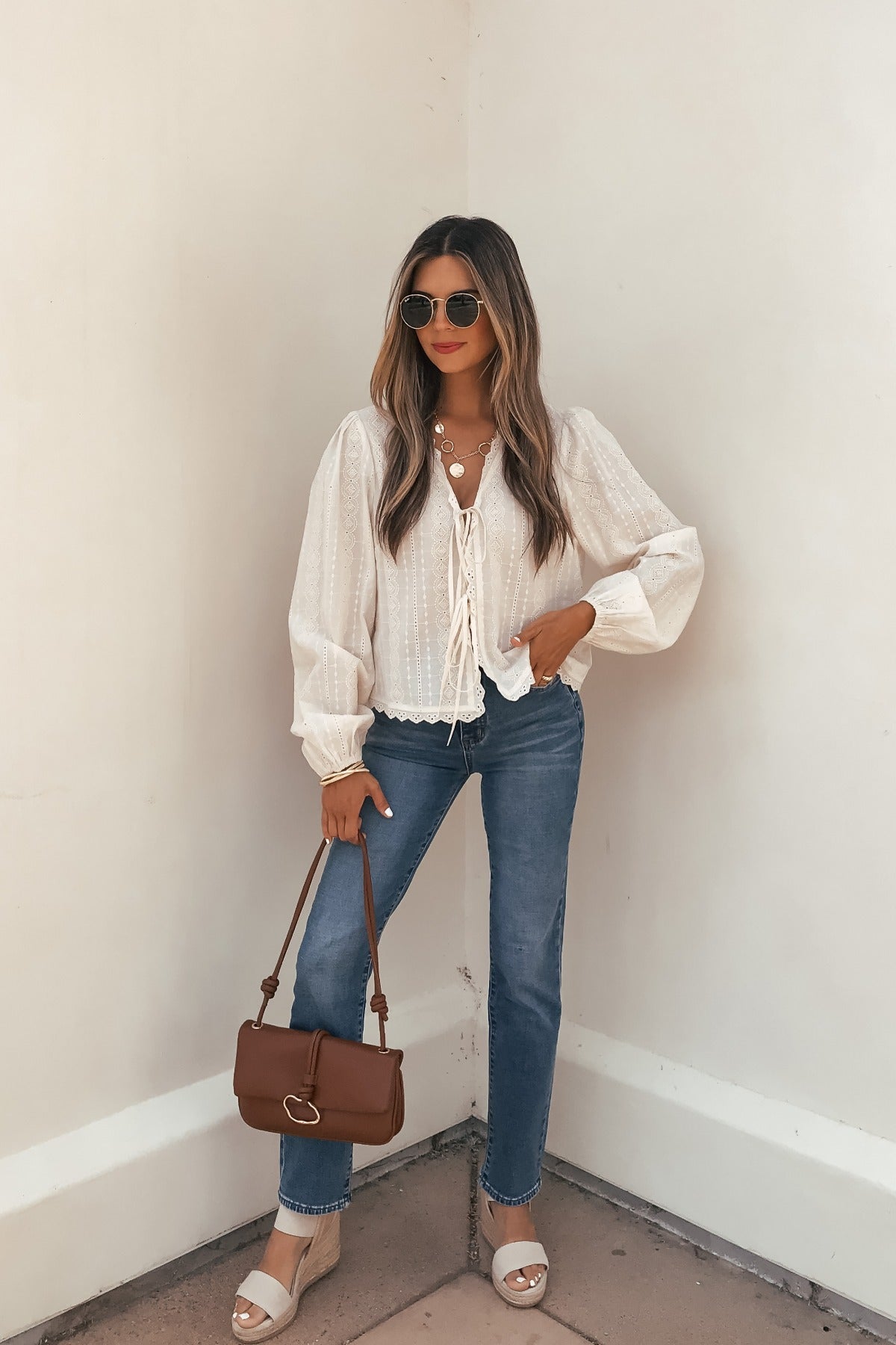 A woman in a Cream Eyelet Tie Front Top, blue jeans, tan sandals, sunglasses, and a brown purse stands by a white wall.