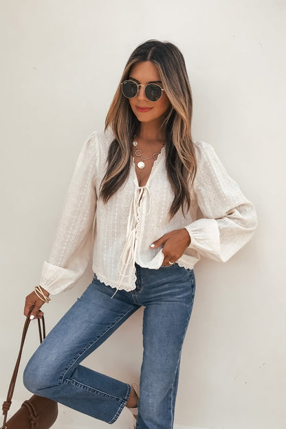 A woman in the Cream Eyelet Tie Front Top, sunglasses, blue jeans, and carrying a brown handbag stands by a white wall.