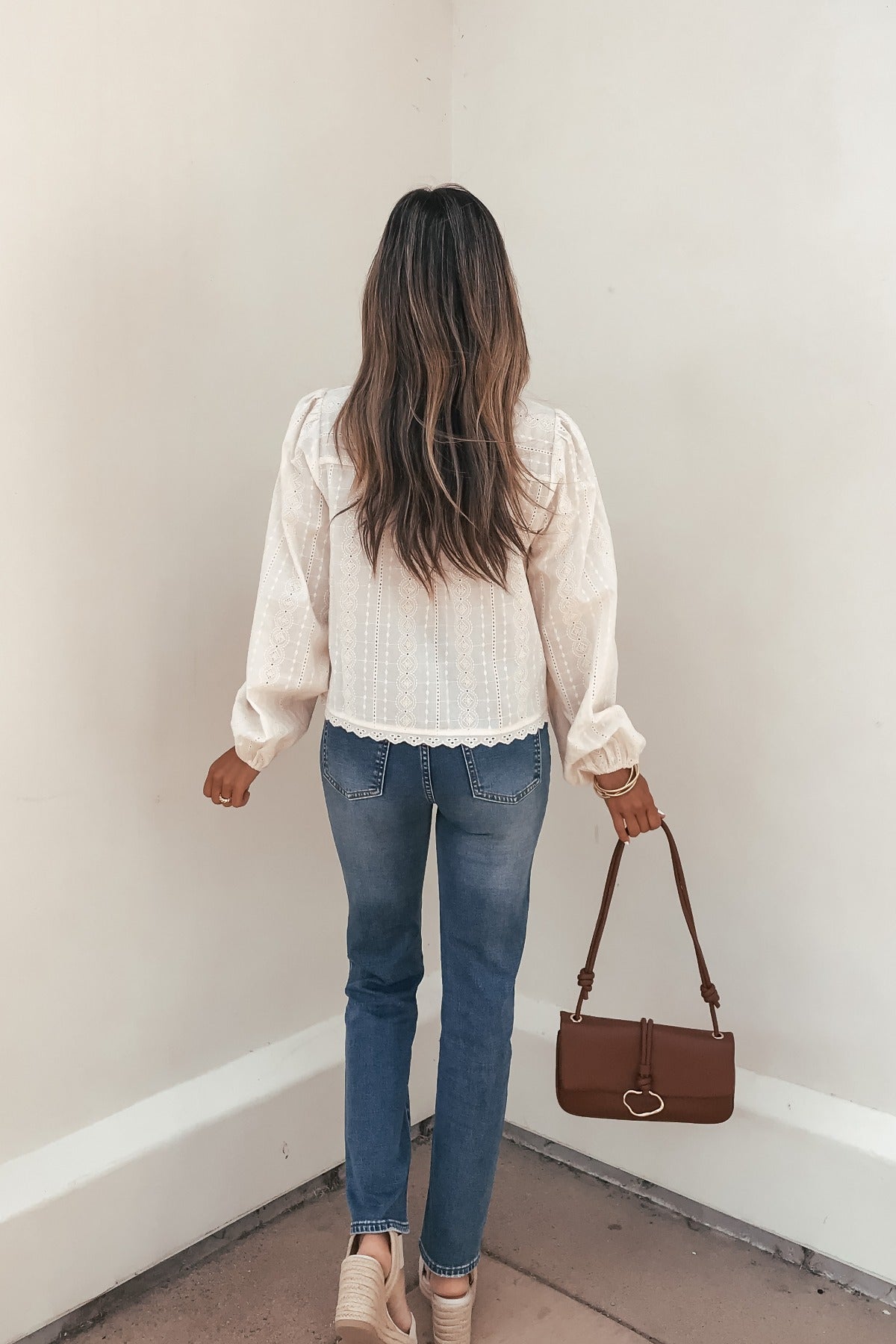 Woman with long, wavy hair wears a Cream Eyelet Tie Front Top, blue jeans, beige shoes, and carries a brown handbag.