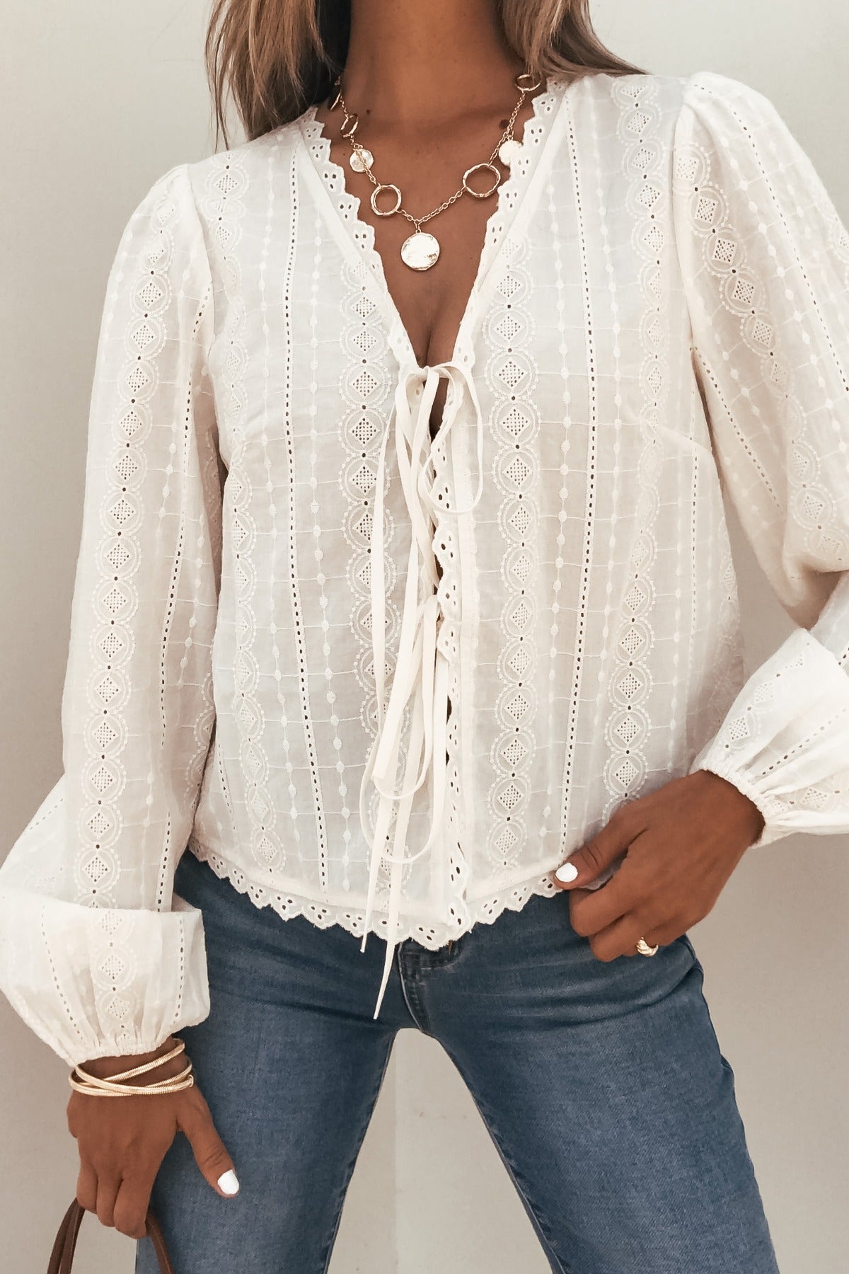 Woman in Cream Eyelet Tie Front Top, blue jeans, layered necklaces & bracelets, standing against a plain background.
