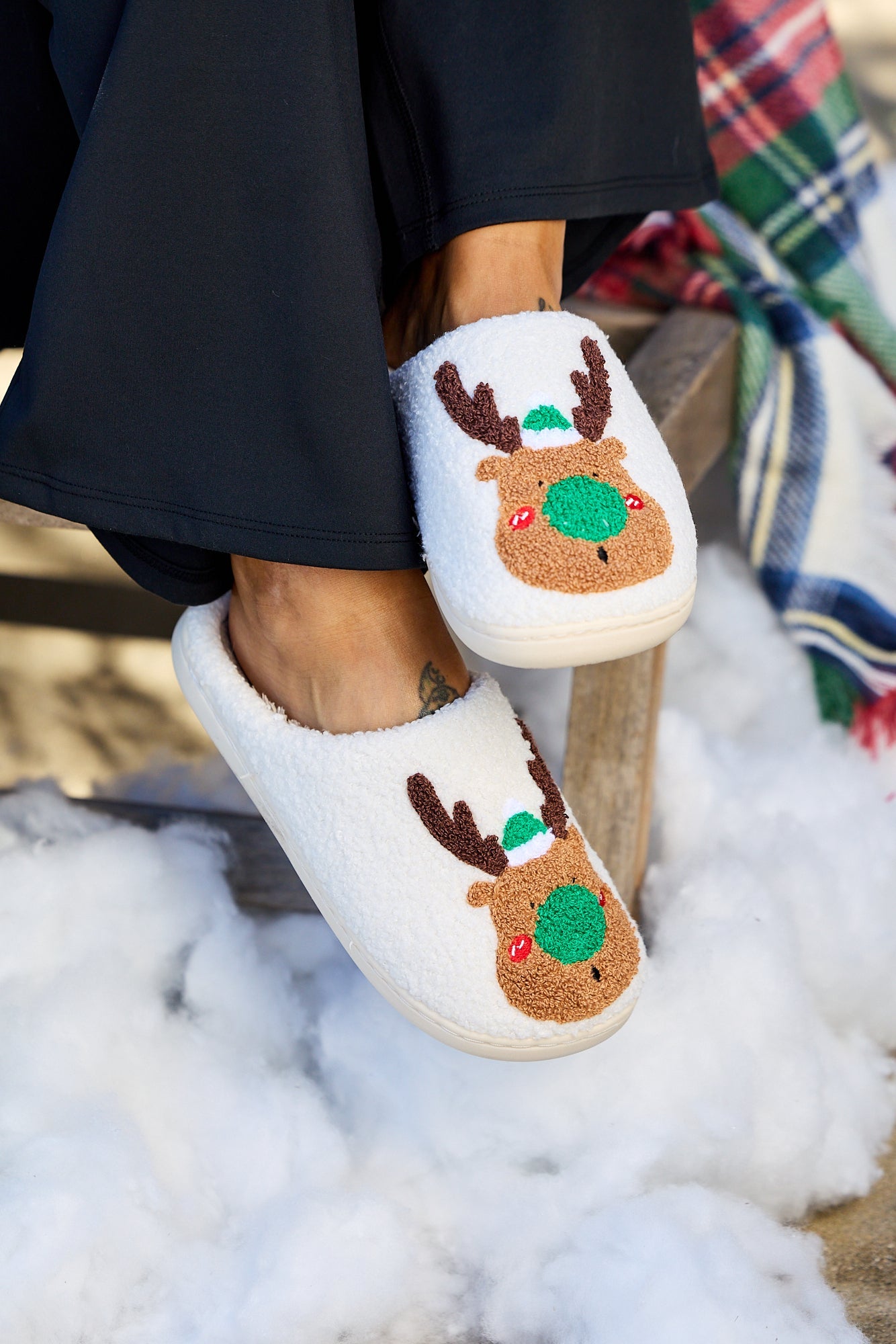 Someone wears Cream Faux Plush Reindeer Slippers, sitting on a wooden bench amid fake snow and a plaid blanket.